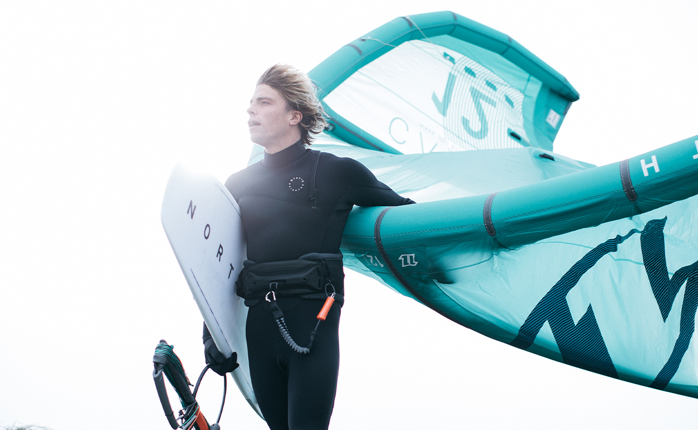 Kitesurfeur en combinaison noire tenant une planche blanche et une aile gonflable turquoise sur fond blanc