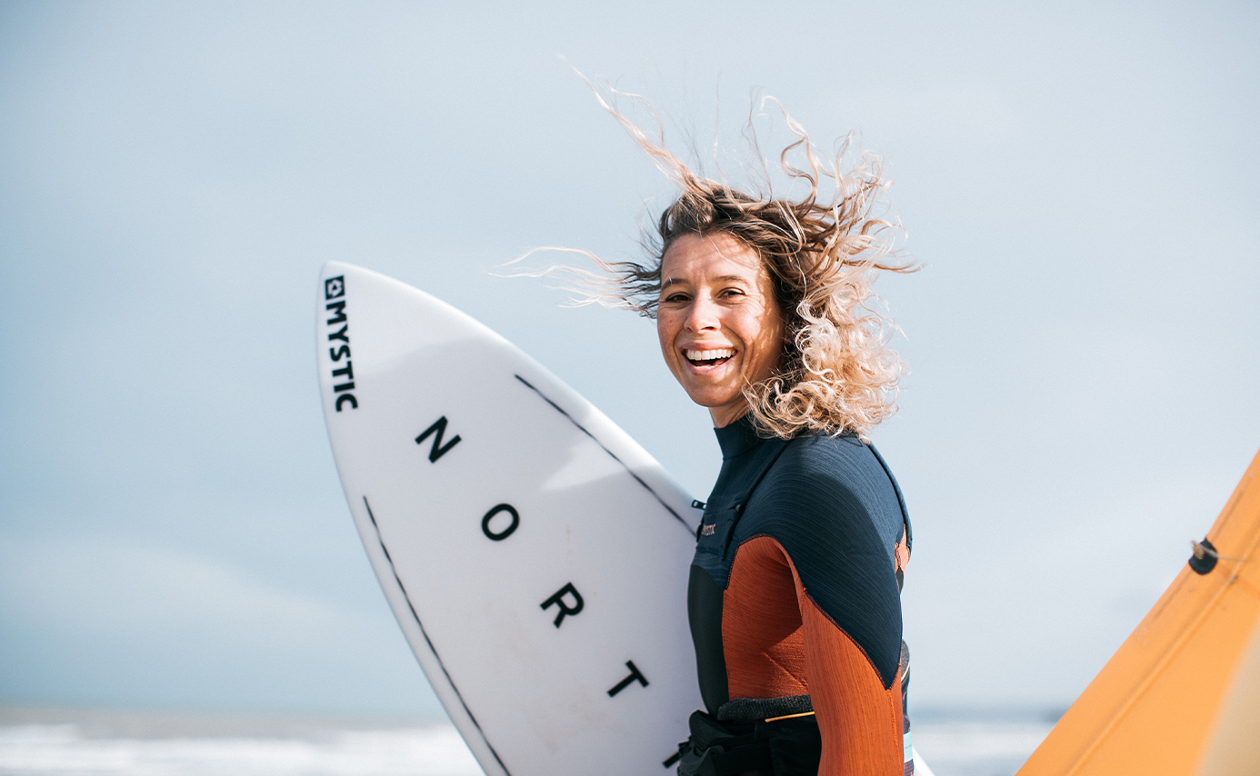 Une femme souriante, cheveux au vent, porte une planche North blanche, en combinaison et harnais orange